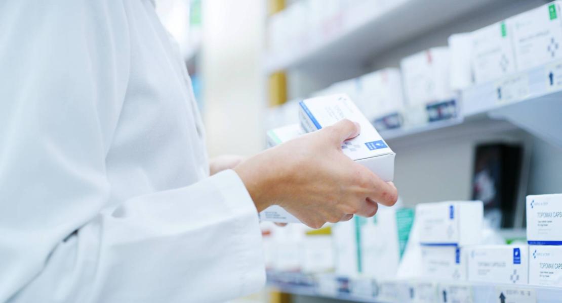 Pharmacist holding a box of medication in front of a shelf stocked with various pharmaceutical products.