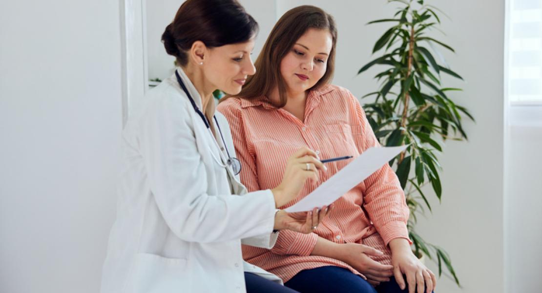 A doctor in a white coat is sitting next to a patient, holding and pointing at a document. The patient, wearing an orange shirt, is attentively looking at the document. They are seated in a well-lit room with a plant in the background.