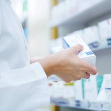 Pharmacist holding a box of medication in front of a shelf stocked with various pharmaceutical products.