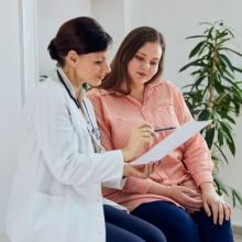 A doctor in a white coat is sitting next to a patient, holding and pointing at a document. The patient, wearing an orange shirt, is attentively looking at the document. They are seated in a well-lit room with a plant in the background.